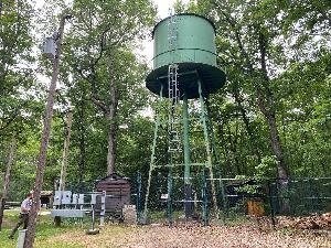 Photo of the green metal Oak Ridge water tower, in between two brown wooden sheds, all of which are surrounded by a chain link fence.