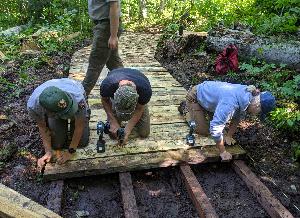 Three park staff attaching wood planks to create a wooden walkway through a muddy section of forest trail
