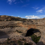 Sipapu Bridge in Natural Bridges National Monument as viewed from above. Photo shows the tan slickrock of the natural bridge with a green ribbon of trees underneath and clear blue skies overhead.