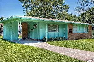 This photo of the Medgar and Myrlie Evers home shows a one-story Ranch-style single-family home that features aqua-colored painted siding and a blonde-brick facade. A one-car carport, the place where Medgar Evers was assassinated in 1963, extends from one side of the house.