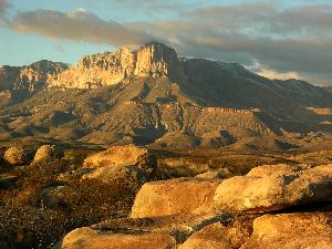 Photo of exposed limestone rock of El Capitan mountain on a partially cloudy day with light from the sunset causing the rocks in foreground to glow a golden yellow.