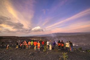 Image of a morning sunrise with long lines of clouds including puffy clouds near the Halema'uma'u crater, blue-grey plume of volcanic smoke rising out of the crater, people standing by a rope line looking at the lava lake in the crater with red lava visible.