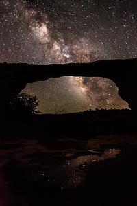 Night sky photo of Owachomo Bridge at Natural Bridges National Monument with the Milky Way and starry sky in the background and star reflections in a small pool in the foreground.