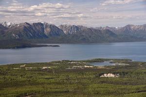 An aerial image of Port Alsworth on the shore of Lake Clark, with trees in the foreground, the town in the mid ground, a blue lake and large mountains behind the town.
