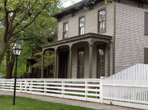 This color photo shows Julius Rosenwald's boyhood home.  There is a white fence and sidewalk with a lamppost in the front of the photo.  A two story home sites closely behind the fence.  There is a porch on the front of the home with 4 pillars.  The first floor has long large windows on the front.  There is a tree visible in the photo and a lush green grass contrasting the grayish home.