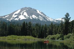 A person paddles a red kayak across Manzanita Lake with a view of evergreen trees and a snow dappled mountain in the background.