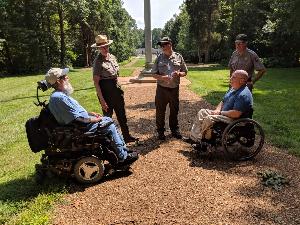 Three park rangers and two men in wheelchairs on a trail. In the background, two parallel trails with a field of grass between the trails and trees on the left and right lead to a parking lot and large stone building.