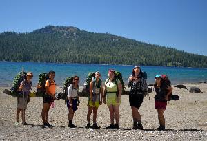 Seven young women wearing backpacking packs pose for a photo at the edge of a lake backed by a conifer-covered peak.