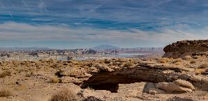 Photograph of reddish-brown sandstone arch known as Skylight Arch. Navajo Mountain and Lake Powell are in the background and blue skies are above. The ground is spotted with small desert bushes.