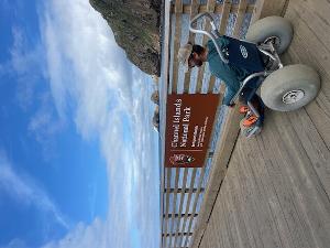 Person sitting in a beach wheelchair on a pier with a sign of Scorpion Ranch, Santa Cruz Island. Cliff and ocean appear in the background.