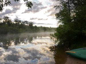 It is early morning on the Namekagon River. Forest frames the river as it curves from the left of the image, around a bend and away from the viewer to the right. Pastel pink and purple clouds in the sky are reflected in the calm, mirror-like water below. A light mist is rising from the water's surface. The bow of a canoe resting on the sandy shoreline can be seen in the lower right corner of the image.