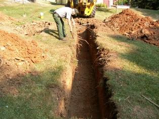 Photograph of trench, which is located within lawn area of residence.  Backhoe and NPS employee pictured.