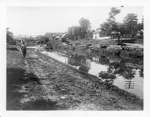 Black and white photograph, circa 1900, showing the Chesapeake and Ohio Canal operation in Hancock, MD.  Canal Company boat is being towed.  Various warehouses visible in photo.  Original iron bridge, crossing the canal is in background.