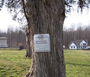 A line of cedar trees at Harris Farm, Spotsylvania Courthouse battlefield, Virginia.  A battle monument at left midground. Recently-constructed estate home at right background.  A rectagular metal sign tacked to the nearest tree reads: "APCWS--Help us protect this battlefield."