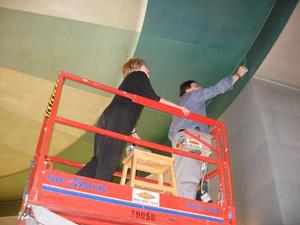 Two park conservators, standing in a sissor lift, conducting tests on paint on the ceiling of the prismatarium.