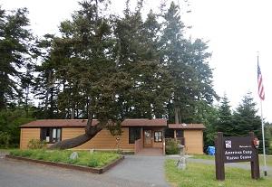 Contemporary photograph of the front of the existing visitor center building. The structure is a prefabricated structure with plain yellowish siding, with few windows, and a gabled asphalt shingle roof. Bathrooms are in a separate matching building just right (east) of the main building and are connected by a walkway.