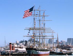 Photo of the park's Hyde Street Pier and historic vessels (Balclutha, with a large American flag flying from her mizzen mast, in the center) looking toward San Francisco from the Bay (city skyline in the background)