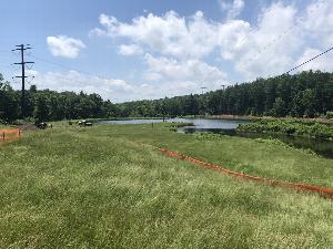 The photo shows the Watergate site prior to construction.  Green grass is in the foreground, and is bisected by orange construction fence.  Barely visible are a truck, a yellow skid-steer, and two people.  In the middleground is a pond, which reflects the blue sky and clouds.  In the background is green forest and above that rises a large transmission line, then the sky.