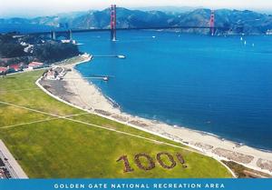 Aerial photo displaying the west end of Crissy Field, the adjacent San Francisco Bay, and the Golden Gate Bridge, with the Marin Headlands behind it. A group of park visitors are arranged on the Crissy Field lawn to spell out "100!".