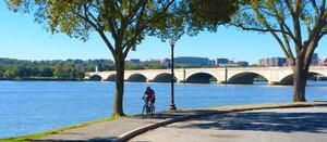 photo of a man on a bike on paved trail next to the Potomac River with Arlington Memorial Bridge in the background.