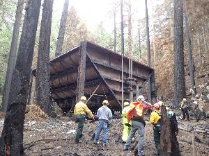 Photo showing the Ladder Creek Settling Tank, which is a large inverted A-frame steel structure supported by four steel I-Beams on each corner.  The tank is is rusted and blackened from a 2015 wildire.  Charred tree trunks and bare soil surround the tank in this photo along with workers in bright colored jackets and shirts and yellow hard hats.