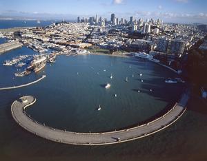 Aerial view of the Aquatic Park Cove showing anchored boats. The curved Aquatic Park Pier is in the foreground, Hyde Street Pier and the Park's historic fleet is at left, the Maritime Museum is at right, and the San Francisco city skyline is in the background.