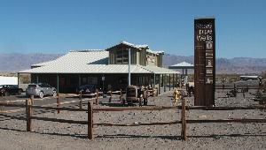 This is a photograph of the Stovepipe Wells General Store. In the foreground, there is a wooden fence surrounding the Stovepipe Wells sign, old machinery, and some vegetation. The General Store and visitors' vehicles are in the mid-ground, and mountains can be seen in the background.