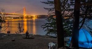 Night-time view of the Golden Gate and lit Golden Gate Bridge from campsite one at Kirby Cove campground, showing a lit blue dome tent partially hidden by trees and a solitary figure seated at a picnic table enjoying the view.