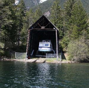 Photo of the Diablo Dry Dock with a boat (the Alice Ross III) parked inside of it.  The Dry Dock consists of rails that slope into Diablo Lake that have a carriage on which the boat is loaded to bring it out of the water.  The upland rails are covered by a large walled structure with a steep pitched roof.  The structure and carriage have since been demolished and removed from the site.
