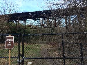 This is a picture of the Foundry Branch streetcar bridge. The view is just north of Canal Road NW, facing north. In the picture, the center trestle section of the bridge is visible, as are several supporting towers on the east side of the bridge. Trees and other vegetation are also visible in front of and behind the bridge. There is a fence in the foreground of the photo with gates chain locked closed. A sign, which reads "Temporary Trail Closure for Bridge Repair", is visible in the foreground to the left.;