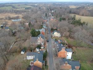 Aerial view of Main Street, Waterford, Virginia. Looking west toward the intersection with Water Street and Second Street. Brick and frame buildings line either side of the paved street. Open green space appear behind the buildings and hills in the distance. (University of Delaware, Center for Historic Architecture & Design, 2018).