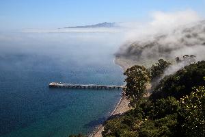 Photo Credit: Tim Hauf
Small bay with calm water, pier, and rocky beach. Fog covers the hillside and mountains are seen in the background.