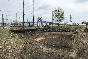 View of an archeological investigation in progress at the site of one of the homes burned in the 1908 Springfield Race Riot. Brick foundations are visible among delineation strings and dark brown dirt in the square dig site, about a foot deep. In the background is the 10th Street Rail Corridor and train tracks, chain link fence, and utility poles.