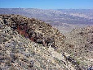 Photo of existing conditions at Skidoo Mill Site. The wooden mill is visible on a steep canyon wall with tailings and remnants of tanks below.