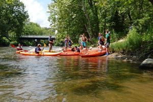 Visitors in brightly colored life jackets board multiple orange, yellow, and red kayaks on the shore of the Cuyahoga River in summer.