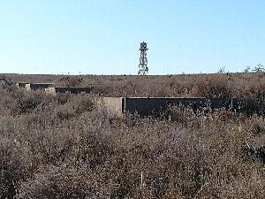 Concrete barracks foundations step down a slight slope in dry grass with the reconstructed Amache water tower rising in the background.