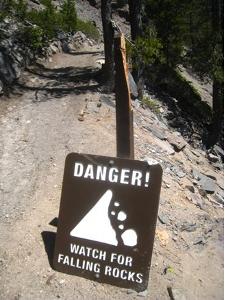 Image of sign with text "Danger! Watch for Falling Rocks" along the Cleetwood Trail in Crater Lake. The sign has been damaged by a falling rock. The sign post is split and the sign is propped up against the broken post.