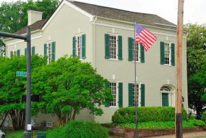 Photo of the Polk home, a two-story light yellow structure with green storm shutters.  The home is surrounded by trees and shrubs.  To the left of the home is a traffic sign showing the home is located on West 7th Street.  A lightpole and an flagpole with an American flag is in the foreground.