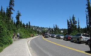 Cars drive, and people walk, the approach to Paradise and Jackson Visitor Center. Cars parked along the roadway on the approach.