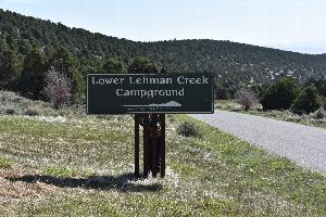Photo of the brown Lower Lehman Creek Campground Sign, adjacent to a paved road and in front of hills with green trees.