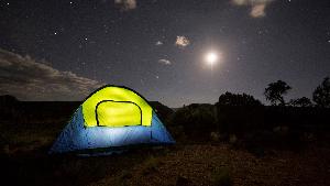 Stars and the moon are visible in a dark night over the desert with the outline of a butte in the background. A glowing green and blue tent is lit from within in the foreground.