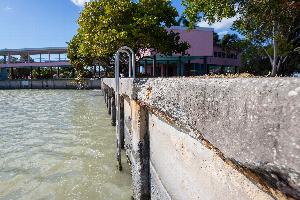 Photo of deteriorating seawall at Flamingo visitor center. Grey concrete cap has spalled due to expansion of rusted reinforcing rod. The visitor center is pink, surrounded by green trees and blue sky. The water of Florida Bay is green.